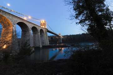 Beautiful night-time view of the historic Menai Suspension Bridge spanning the Menai Strait illuminated at dark, Isle of Anglesey, North Wales