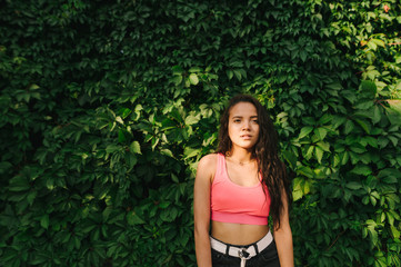 Attractive curly hispanic girl in stylish casual clothes stands against a background of green wall with ivy. Fashionable street girl in sportswear on wall background with green vine.