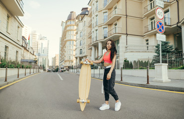 Attractive girl in casual clothes posing on city streets with a longboard in her hands, wearing...
