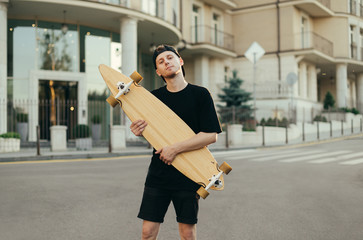 Handsome teenager in casual clothes and a cap stands with a longboard in his hands on the street and looks in camera with a smile on his face. Guy skateboarder posing with a longboard in his hands © bodnarphoto