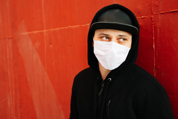 a young man in black sportswear and a baseball cap with a medical mask on his face near the red wall.