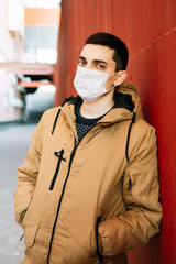 a young man in a brown jacket with a medical mask on his face on the street, near the red wall.