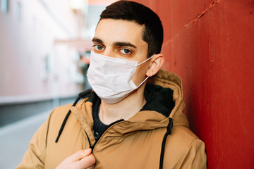 a young man in a brown jacket with a medical mask on his face on the street, near the red wall.