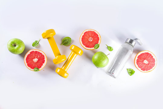 Fitness and healthy food lifestyle concept. Dumbbells, diet fruit and vegetable lunch box, water and jump rope on white background. Flatlay image, top view copy space