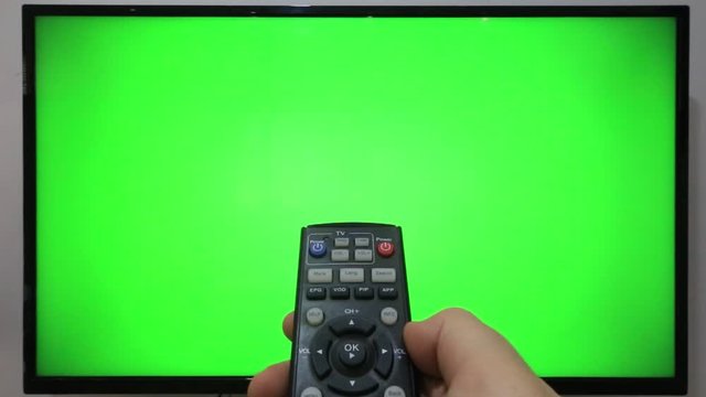 Person pointing a TV remote and pressing buttons in front of a TV with green screen