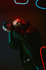 Fototapeta na wymiar bi-racial cyberpunk player in protective mask aiming gun near neon lighting on black