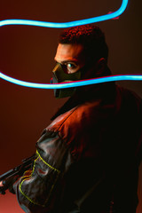 selective focus of bi-racial cyberpunk player in protective mask holding gun near blue neon...