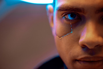 cropped view of mixed race cyberpunk player with metallic plates on face and blue eyes looking at...