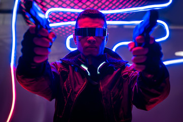selective focus of armed mixed race cyberpunk player in futuristic glasses holding guns near neon...