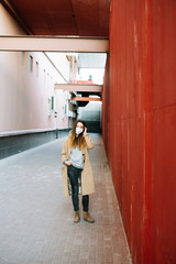 a young girl in a brown coat with a medical mask on her face on the street, near the red wall.