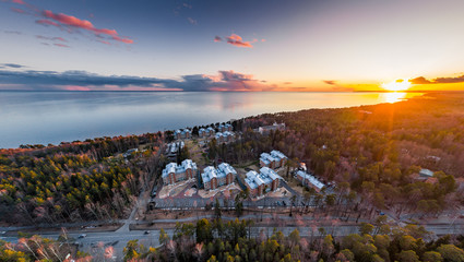Roofs of houses in the forest, the picturesque coast at sunset, quiet water, tops of trees are illuminated by the sunset sun, pink clouds