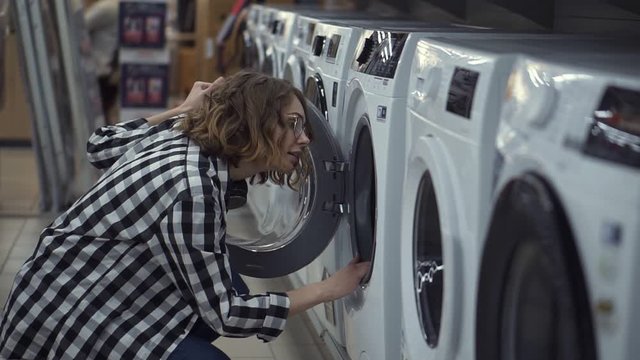 A young positive woman in a plaid shirt choosing washing machine in the shop of household appliances. Opens the door and look inside. Side view. Slow motion
