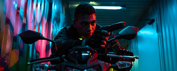panoramic shot of bi-racial cyberpunk player on motorcycle aiming gun on street with graffiti
