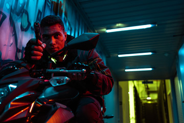Fototapeta na wymiar dangerous and mixed race cyberpunk player on motorcycle aiming gun on street with graffiti