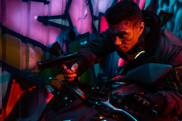Fototapeta na wymiar mixed race cyberpunk player on motorcycle aiming gun on street with graffiti
