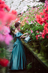 Obraz na płótnie Canvas Beautiful romantic young woman in a greenhouse with azaleas. The girl is holding a blue watering can. Art portrait of a girl wearing blue magnificent vintage dress.