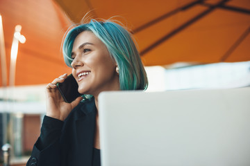 Busy caucasian entrepreneur with blue hair having a phone discussion while resting with a computer in a coffee shop
