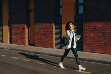 Gorgeous student with curly hair is walking while having a phone discussion and holding laptop