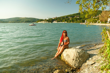 A young woman with African pigtails in a swimsuit sits on a stone on the shore of a mountain lake.