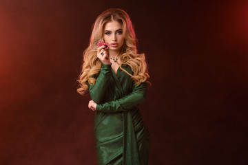 Blonde woman, bright make-up, green stylish dress and jewelry. Showing two red chips, propping her elbow, posing on brown smoky background. Close-up