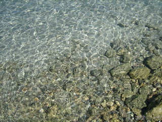Through the clear water in the rays of the reflection of the sun, you can see the shallow sea floor of sand, pebbles and stones of a gray brown color.