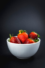 A white bowl with precious strawberries inside with a black background