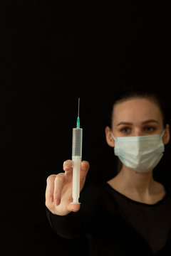 Beautiful girl in a protective mask and with a syringe in her hand. Protect yourself from the virus. Black clothes and background. Protection.