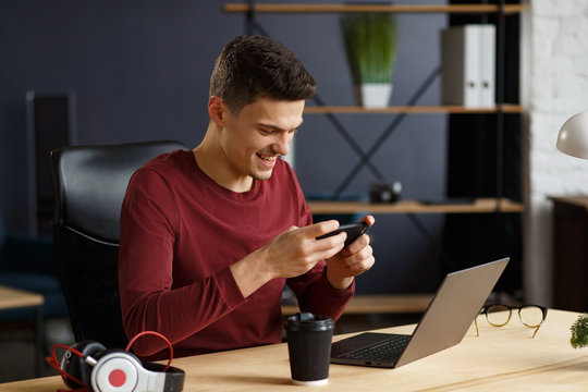 Young Man Playing Games On Mobile Phone In Home Office With Laptop. Relaxation, Taking A Break. Young People Working With Mobile Devices.