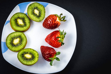 Sliced kiwis and strawberries on a white plate with a black background