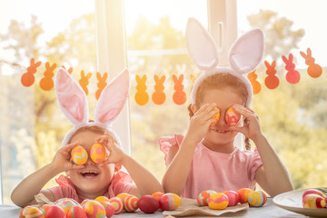 Cute girls holding yellow easter eggs instead of eyes and laughs, having fun on a background of windows. Children celebrate Happy Easter
