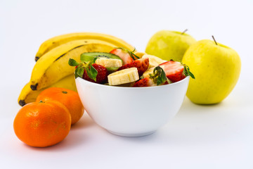 A fruit salad cut in a white bowl on a white background next to tangerines, bananas and apples