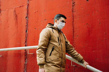 Obraz premium a young man in a brown jacket with a medical mask on his face on the street, near the red wall.
