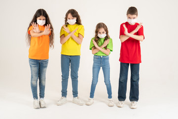 Fototapeta premium Group of schoolchildren children in colorful T-shirts and medical masks showing crossed hands gesture while looking at the camera over white background. Isolated. Stay at home. Say No to the
