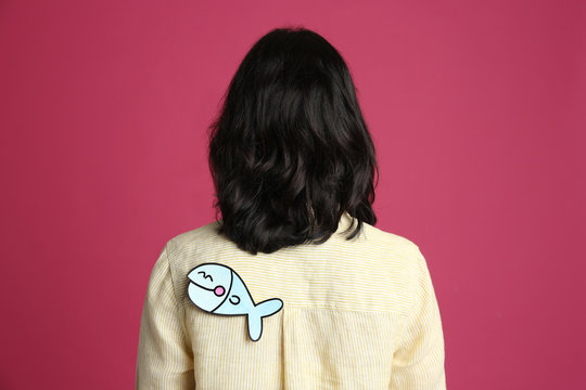 Woman with paper fish on back against pink background. April fool's day