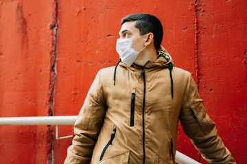 a young man in a brown jacket with a medical mask on his face on the street, near the red wall.