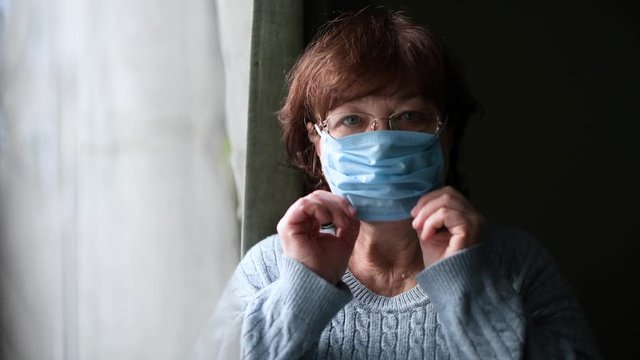 An elderly woman puts on a protective medical mask near the window, looks at the camera, then removes the mask and sighs with relief