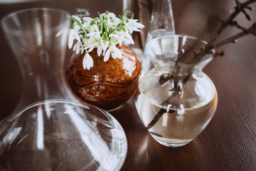 Snowdrops in a clay vase between glass containers on the table. Concept for spring. The first flowers. Still life