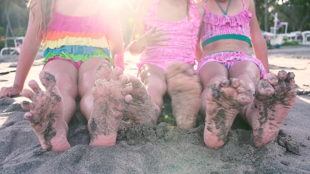 Close up of the feet of three girls sitting on the sand on the beach on a sunny day