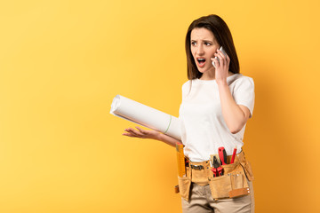 shocked handywoman talking on smartphone and holding project on yellow background