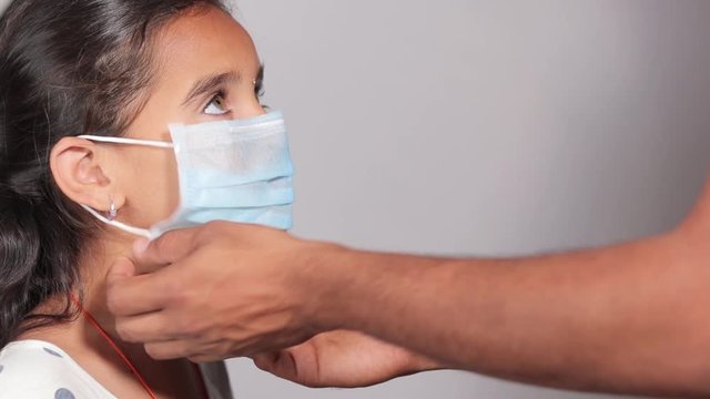 Prevention of spreading the coronavirus or COVID-19 outbreak by wearing mask - Father wearing a disposable hygienic face mask to his young girl daughter to protect spread of disease.