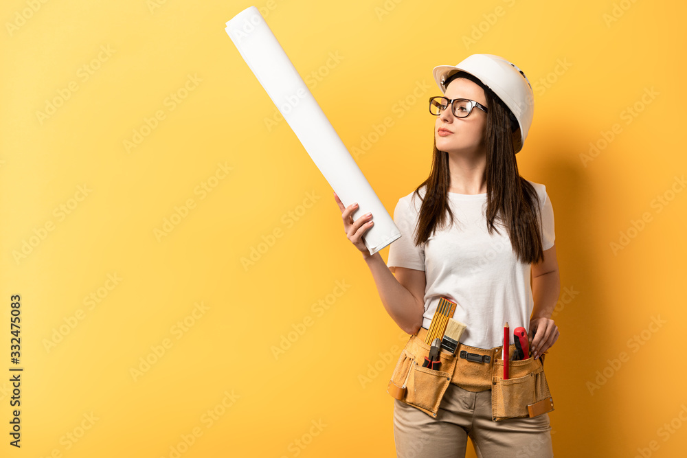 Wall mural thoughtful handywoman holding and looking at blueprint on yellow background