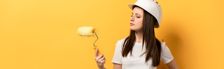panoramic shot of serious handywoman holding paint roller on yellow background