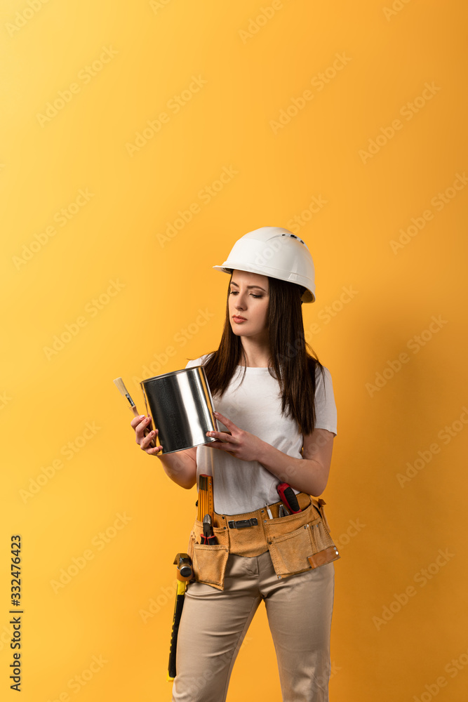 Wall mural serious handywoman holding paint can and paint brush on yellow background