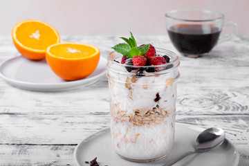 Overnight oats with yogurt topped with berries in a glass jar. Coffee and oranges in the background