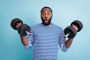 Fototapeta na wymiar Photo of handsome dark skin guy open mouth lifting two heavy dumbbells weight practicing hard gym strong arms wear striped sailor shirt isolated blue color background