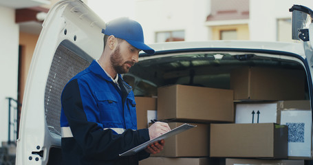 Portrait of the Caucasian good looking male worker of shipping company standing at the van with boxes and writing the document, then smiling to the camera. Outdoor.