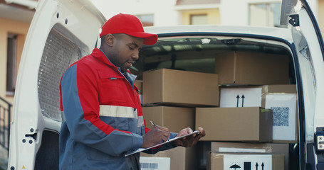 Portrait of the african American young male worker of shipping company standing at the van with boxes and writing the document, then smiling to the camera. Outdoors.