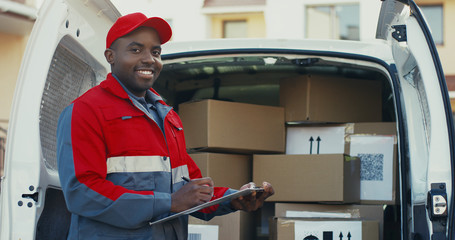 Portrait of the african American young male worker of shipping company standing at the van with boxes and writing the document, then smiling to the camera. Outdoors.