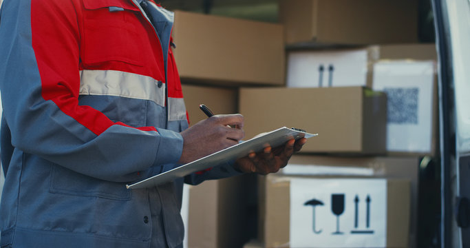 Close up of the African American male hand writing on the clipboard. Deliveryman standing at the van with male boxes. Outside.