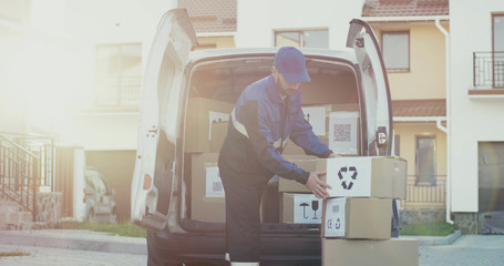 Caucasian male courier in the blue costume and a cap taking out mail carton boxes from the white van on the sunny day in the street. Outdoor.